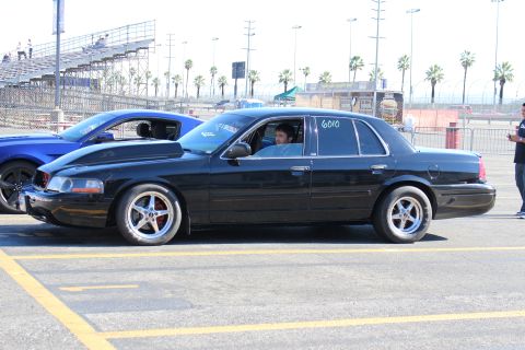 2003 Ford Crown Victoria - Side view of a black full-size sedan numbered 6010 with a driver inside and polished five-spoke wheels at a racetrack staging area.