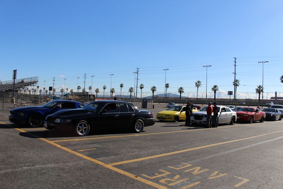 Black sedan and dark coupe parked in a dragstrip lot under clear blue sky with palm trees