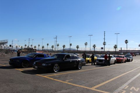 2003 Ford Crown Victoria - Several cars including a blue coupe and a black sedan line up across a sunny dragstrip parking lot beneath palm trees.