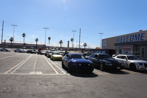 2003 Ford Crown Victoria - blue Mustang, black sedan, and silver Chrysler 300 are parked beside a large building marked WELCOME at a dragstrip.
