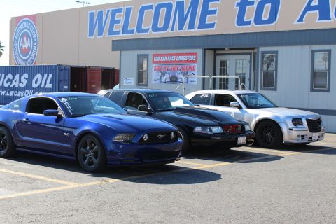2003 Ford Crown Victoria - blue Ford Mustang GT with a 5.0 badge, a black Mustang, and a silver Chrysler 300 parked outside a AAA dragway building.