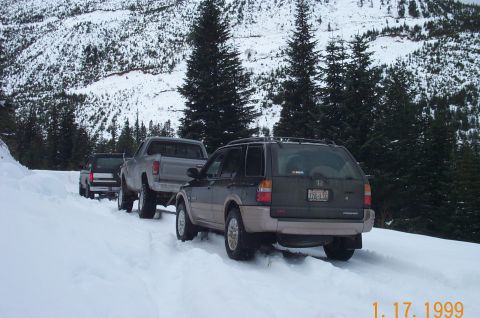 1995 Ford Bronco - Three vehicles navigate a snowy mountain road, with a dark SUV in the foreground and two trucks behind it.