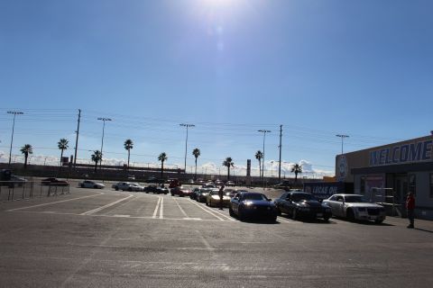 2003 Ford Crown Victoria - wide sunlit dragstrip parking lot with cars parked along a WELCOME building and a person standing on the right.