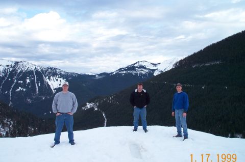 1995 Ford Bronco - Three men pose for a photo on a snowy mountain ridge, with a vast expanse of forested valleys and snow-capped peaks stretching out behind t…