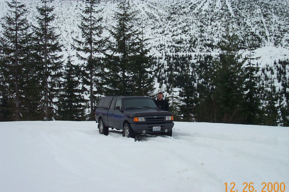 Dark blue Ford Ranger pickup truck with a cap on the bed, parked in the snow with a person standing next to it