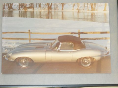 1992 Ford Bronco - A silver convertible Jaguar E-Type is parked on a road with a wooden fence and snow-covered ground in the background.