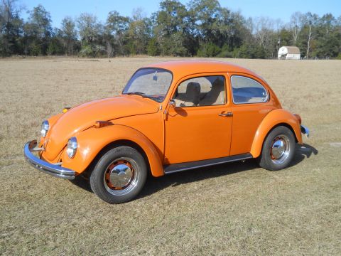 1992 Ford Bronco - A bright orange Volkswagen Beetle is parked on a grassy field with a few trees and a small building in the background.