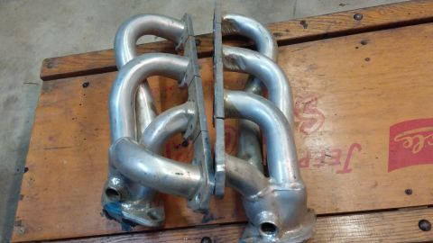 1995 Ford Bronco - A pair of silver exhaust headers are laid out on a wooden surface. The headers are curved and appear to be made of metal, with flanges at t…