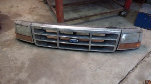 1995 Ford Bronco - A chrome and brown front grille assembly with integrated headlights is displayed on a concrete floor. The Ford logo is visible in the cente…