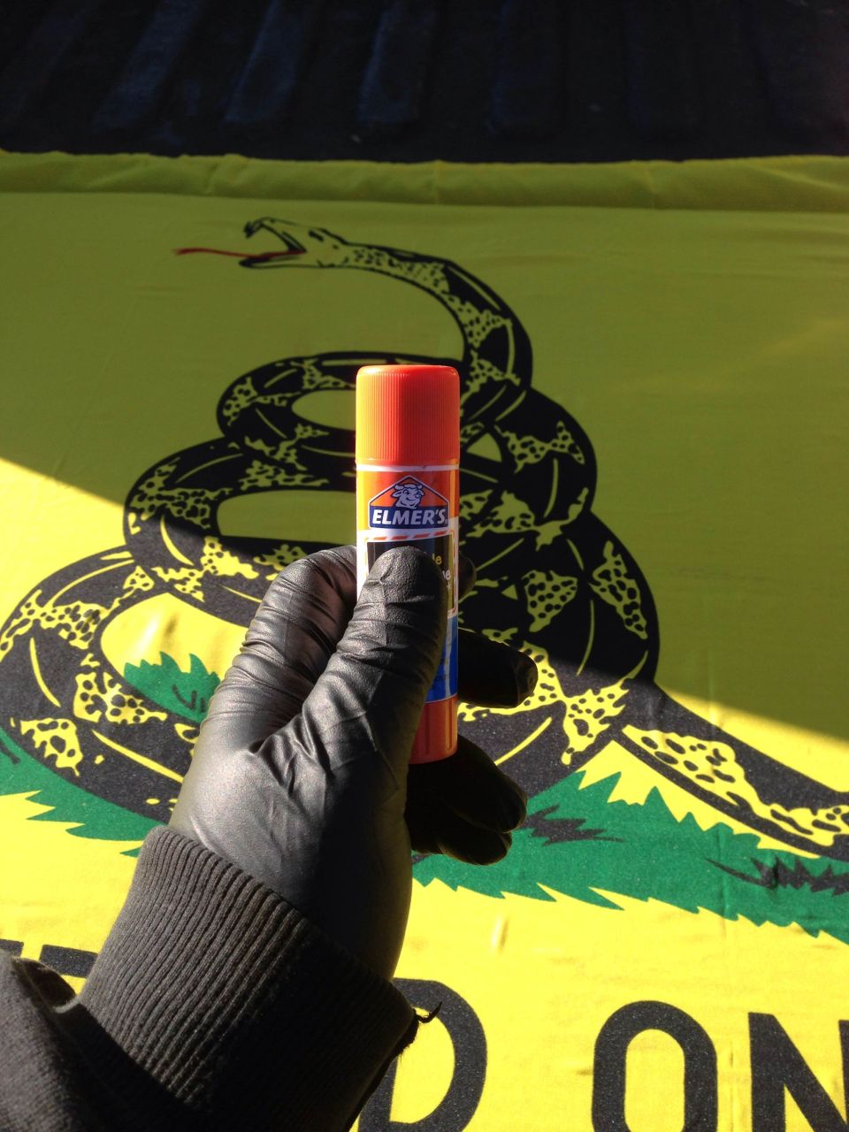 Gloved hand holding Elmer's glue stick in front of a Gadsden flag.