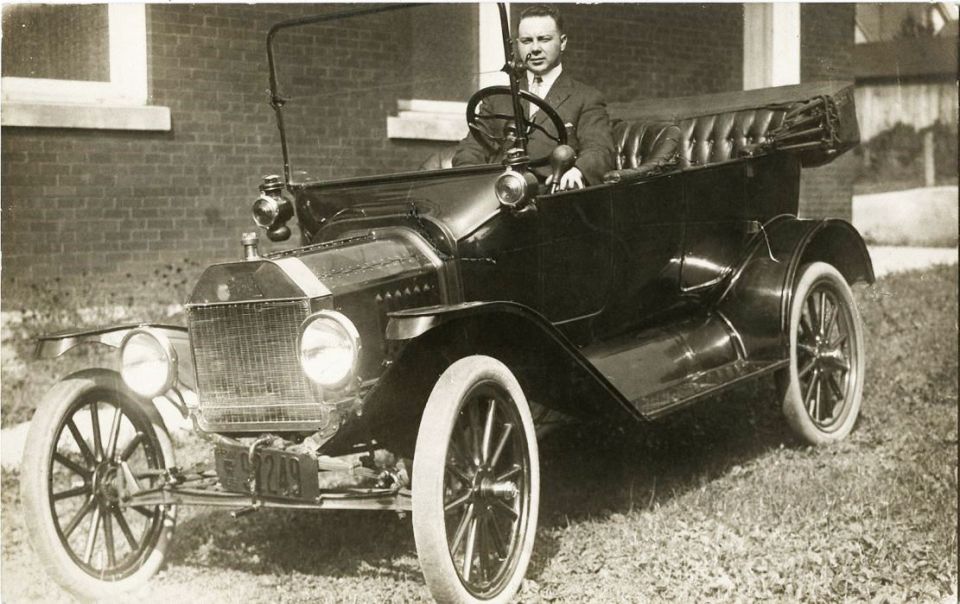 Man in a suit driving a dark touring car parked on grass.