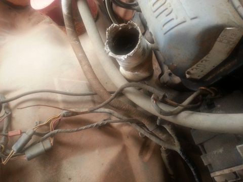 1973 Ford F-100 - A close-up view of a damaged flexible metal exhaust pipe connected to an engine component in a vehicle's engine bay. Various wires and hose…