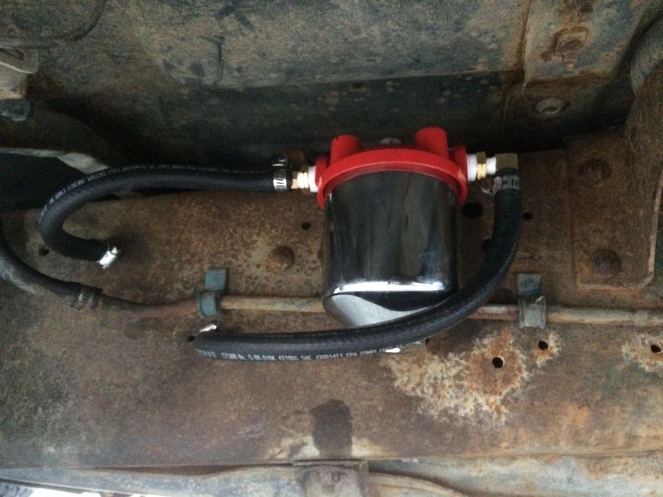 Underneath view of a black fuel filter with red top and black hoses mounted on a rusty vehicle frame.