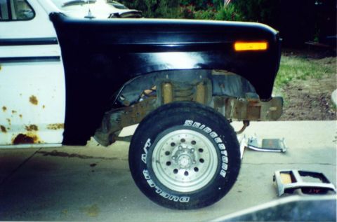 1981 Ford Bronco - The vehicle's front fender and wheel are shown, with rust visible on the lower body panel. Parts are laid out on the ground.