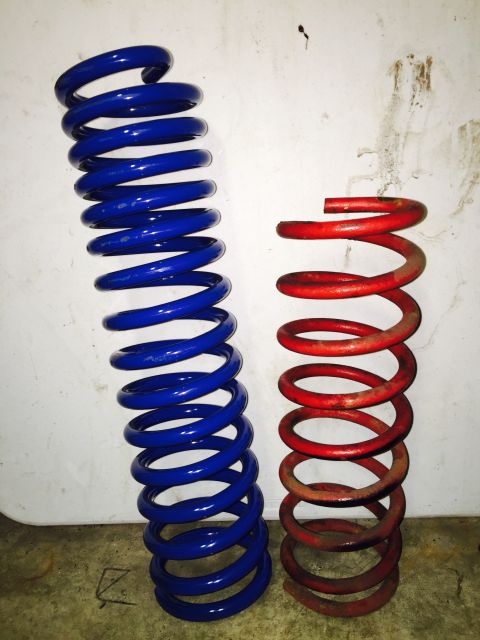 1974 Ford Bronco - Two suspension coil springs are shown standing upright against a white wall. The taller spring on the left is blue, and the shorter spring…