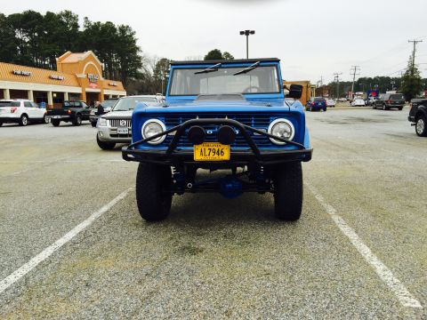 1974 Ford Bronco - The front view of a bright blue classic Ford Bronco is parked in a gravel parking lot. The vehicle has a black brush guard and a yellow Vir…