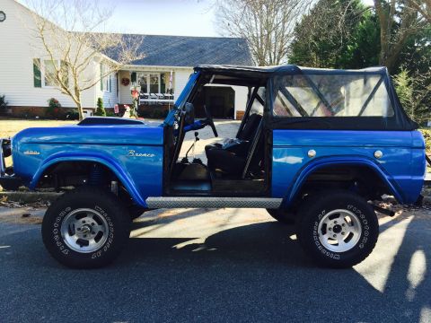 1974 Ford Bronco - A bright blue 1974 Ford Bronco is parked on a residential street, viewed from the side. The vehicle features a soft top, removed doors, and…