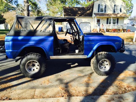 1974 Ford Bronco - A bright blue, lifted Ford Bronco with a soft top is parked on a residential street. The vehicle features large off-road tires and a roll c…