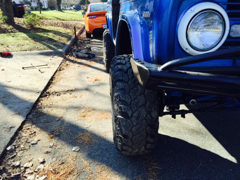 1974 Ford Bronco - A close-up, low-angle shot of the front driver's side wheel and fender of a blue 1974 Ford Bronco. A jack and lug wrench are visible on the…