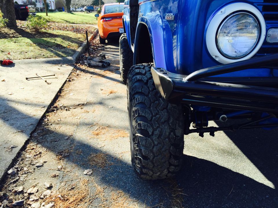 Blue 1974 Ford Bronco front driver's side wheel and fender with jack and lug wrench on sidewalk.