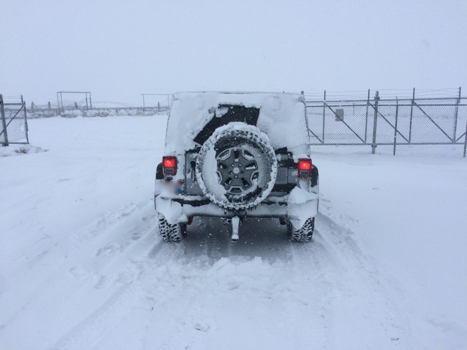 Rear view of a 2014 Jeep Wrangler covered in snow, parked on a snowy road near a fence