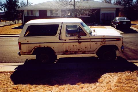 1981 Ford Bronco - A heavily rusted white 1978 Ford Bronco sits parked on a residential street next to another dark-colored car.