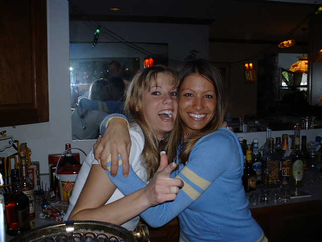 Two women hugging and smiling at a bar with liquor bottles behind them
