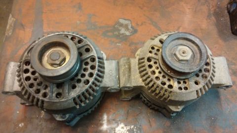 1995 Ford Bronco - Two alternators are mounted side-by-side on a workbench. Each alternator has a pulley attached to the front.