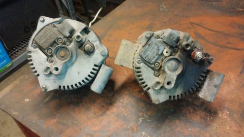 1995 Ford Bronco - Two automotive alternators are shown side-by-side on a workbench. The alternator on the left appears to be a Motorcraft part, while the one…
