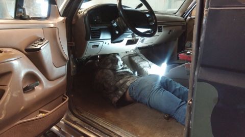 1995 Ford Bronco - A person in jeans and a camouflage hoodie is lying on the floor of a vehicle, working under the dashboard. The interior shows tan door pane…