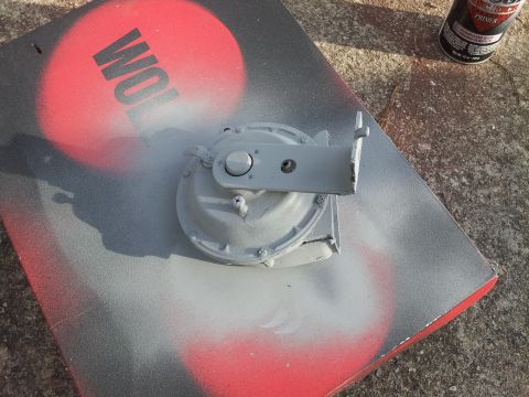 1976 Ford Ranchero - A gray painted part, possibly a horn assembly, sits on a piece of cardboard with red and gray spray paint splatters. A can of primer is vis…
