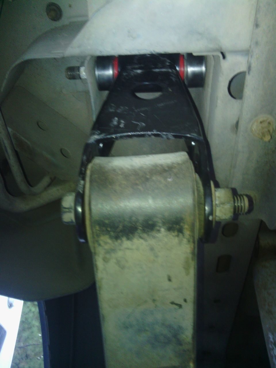 Underside view of vehicle suspension with black metal component, red bushings, and leaf spring.