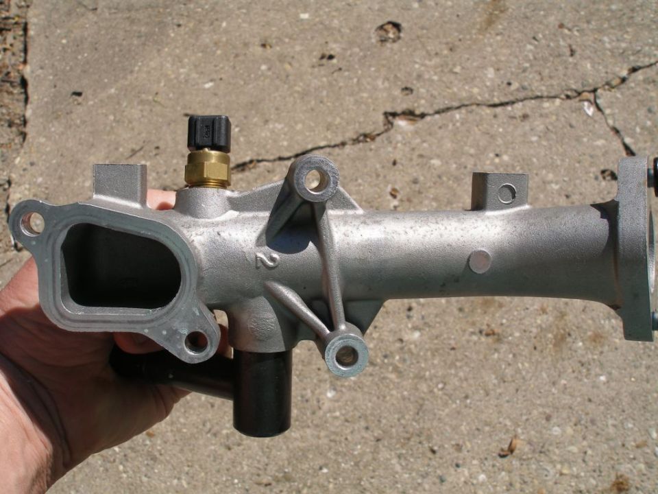 Silver metal automotive thermostat housing with sensor and black hose attachment held by hand