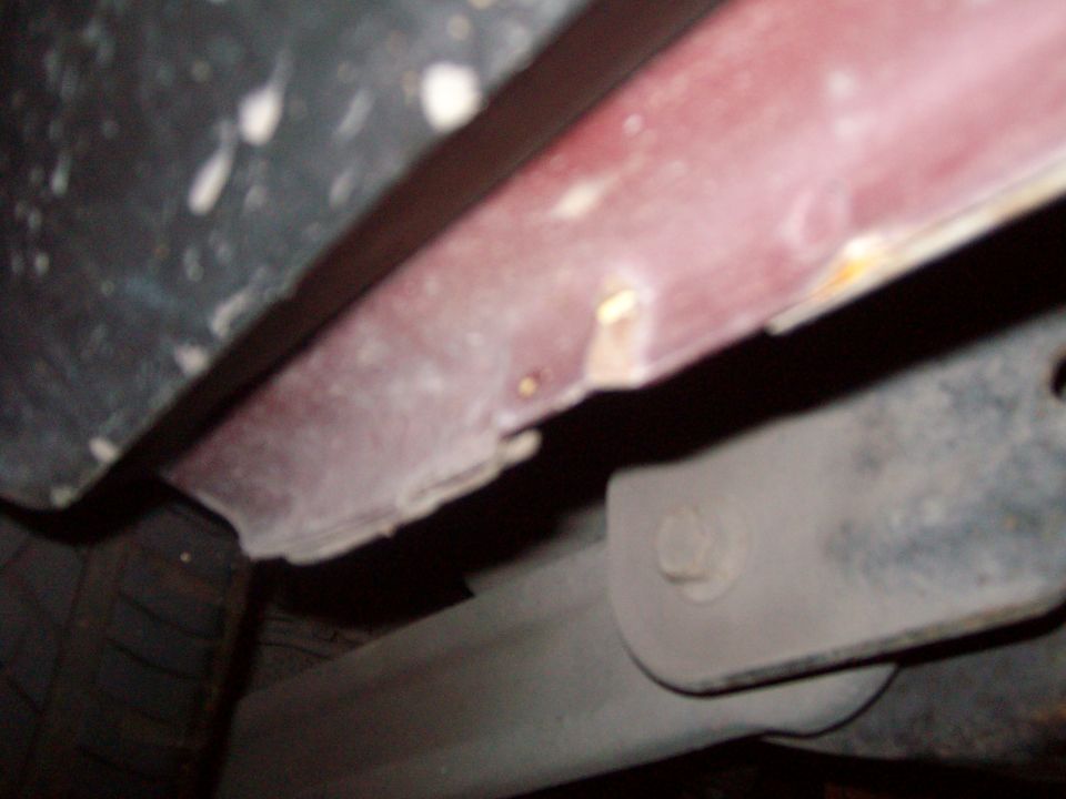 Underside view of a vehicle showing rusted red panel, gray bracket with bolt, and part of a tire.