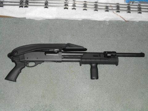 Black tactical shotgun with pistol grip and vertical foregrip on grey carpet, model train tracks in background