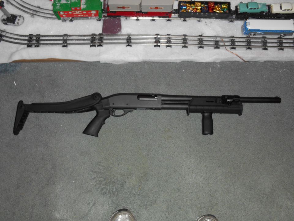 Black shotgun with collapsible stock and foregrip on gray carpet, model train set in background.