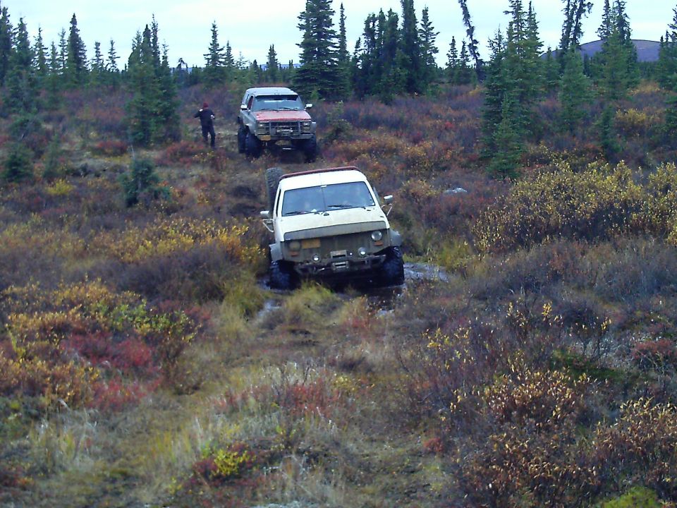 Two off-road vehicles, a red Ford truck and a white SUV, driving through a muddy, brushy landscape.