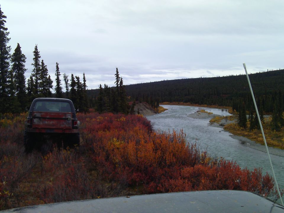 Rear view of a red and black pickup truck parked in a field of autumn brush by a river.