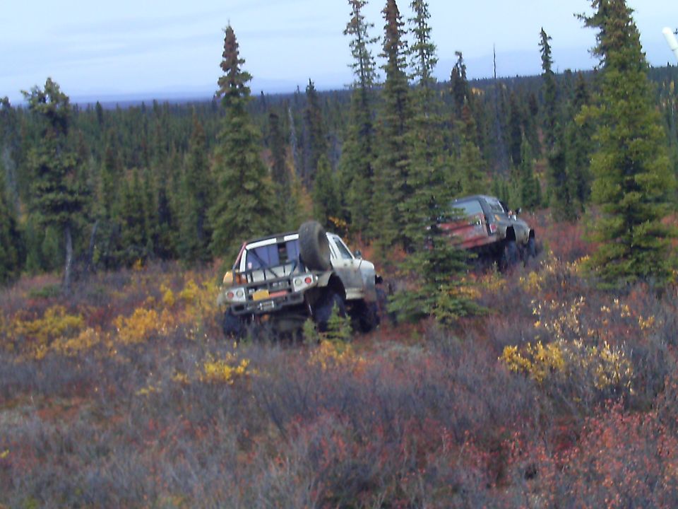 Two off-road pickup trucks driving through a wooded area with autumn colors.