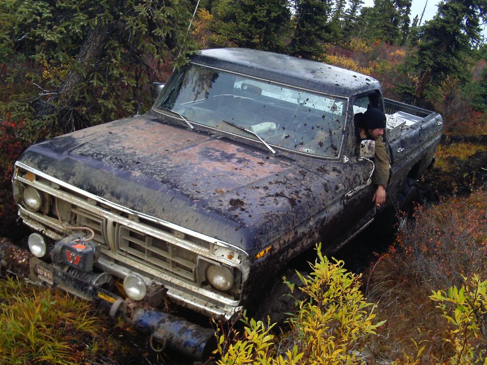 Mud-covered dark pickup truck stuck in woods, driver leaning out window.