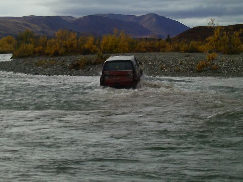 1990 Ford F-250 - A red SUV navigates a river crossing, with autumn foliage and mountains visible on the shore.