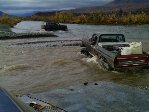 1990 Ford F-250 - Two trucks navigate a shallow river crossing in a remote, scenic landscape with autumn foliage.