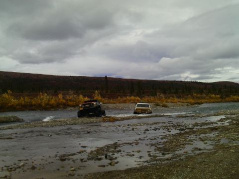 1990 Ford F-250 - Two vehicles navigate a rocky riverbed on an overcast day, with autumn foliage visible on the distant hills.