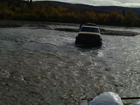 1990 Ford F-250 - A white SUV navigates through a shallow river, with autumn foliage visible on the distant hills.