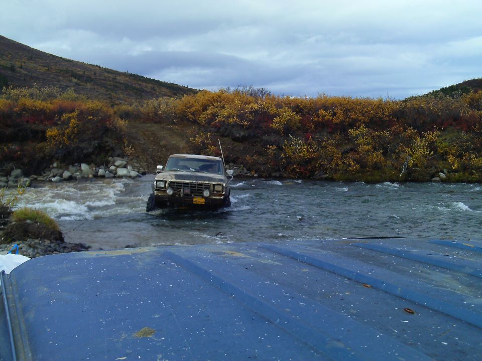 Ford F-Series truck driving through a river with fall foliage on the banks. Another vehicle's blue hood is in the foreground