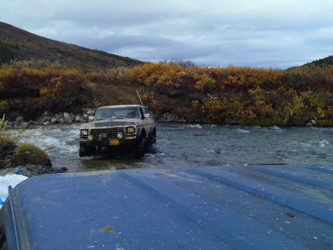 1990 Ford F-250 - A Ford F-Series truck navigates a shallow river with autumn foliage in the background.