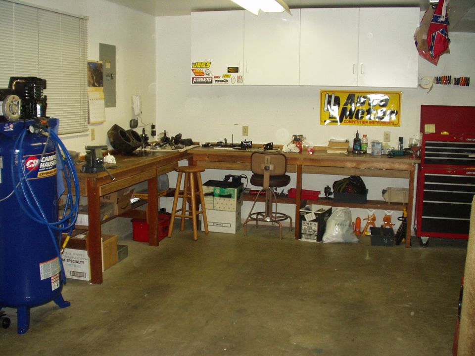 Garage workbench with blue air compressor, red tool chest, and Auto Meter banner; no vehicle shown