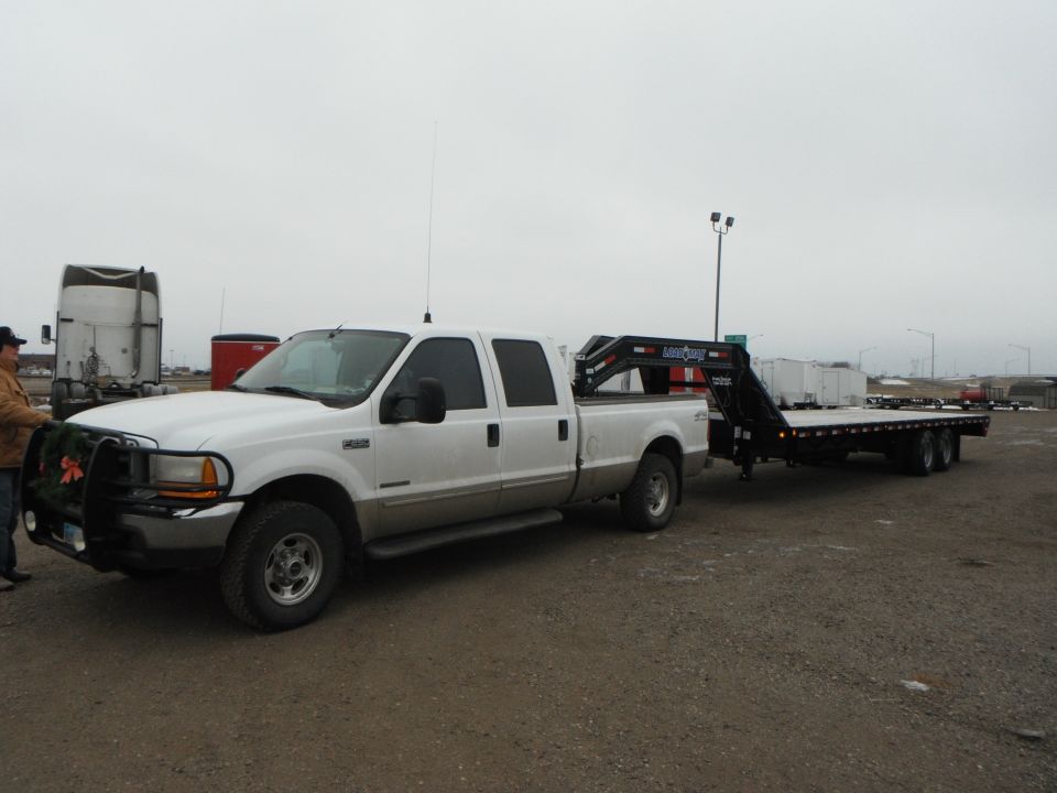 White pickup truck with a gooseneck trailer attached. Truck has a brush guard with a wreath on it