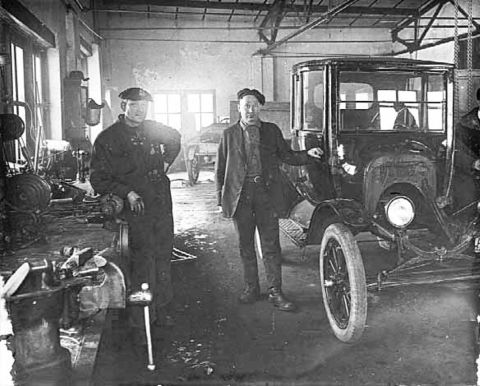 1909 Ford Model T - Two men stand in a garage next to a 1909 Ford Model T, with tools and equipment visible in the background.