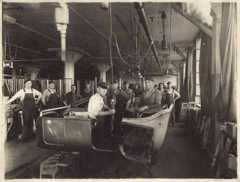 1909 Ford Model T - Workers are busy assembling a car body on the factory floor, with tools and materials visible around them.
